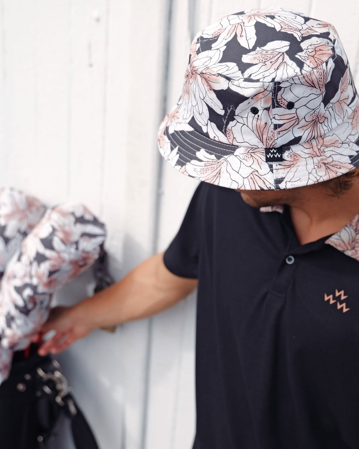 A person wearing a dark bucket hat featuring an azalea print and a matching black polo shirt.