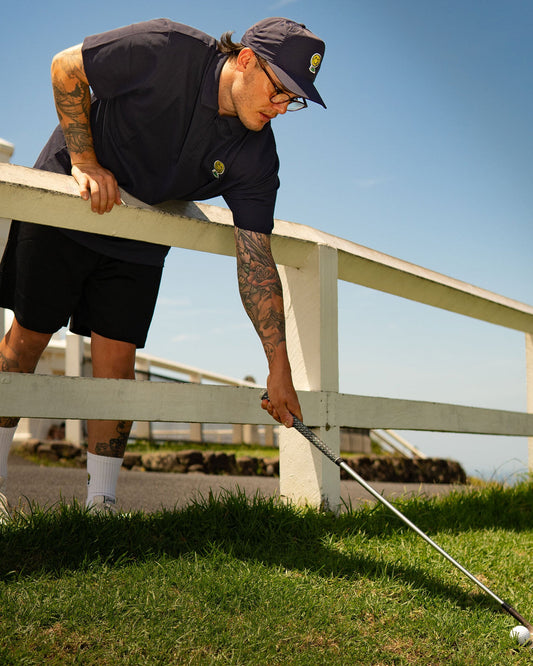 A man wearing a navy quarter-zip polo shirt leans over a white fence to retrieve a golf ball with a club.