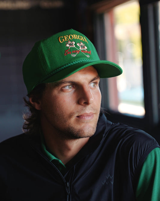 A man wearing a Masters green snapback with a braided brim cord and embroidered text and floral logo.