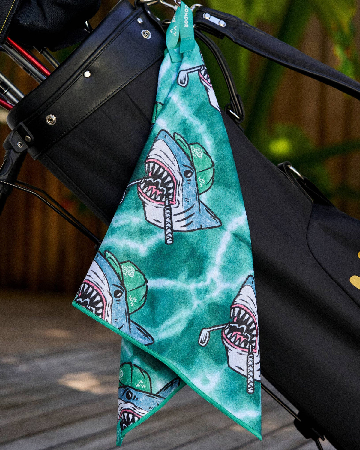 A green tie-dye golf towel hanging from a black golf bag, featuring a printed shark graphic design.