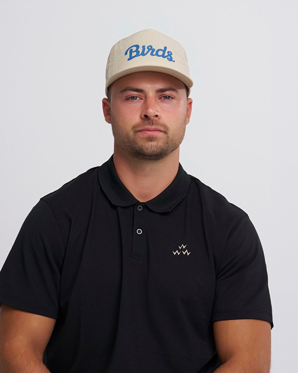 A man wearing a tan snapback cap with a blue script logo and a black polo shirt.