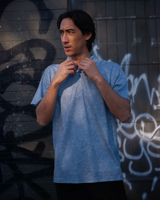 A man wearing an acid blue polo shirt, buttoning the collar against a graffitied wall.
