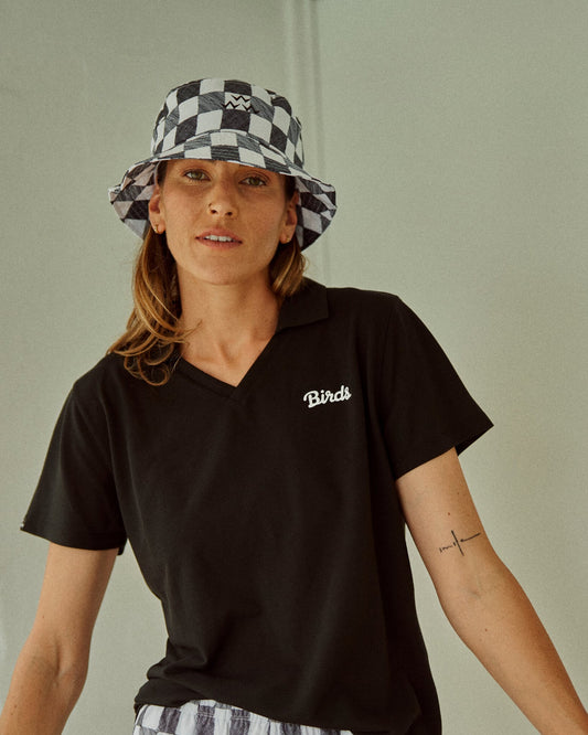 A woman wearing a black and white gingham bucket hat featuring a golf babe logo and a black v-neck shirt.