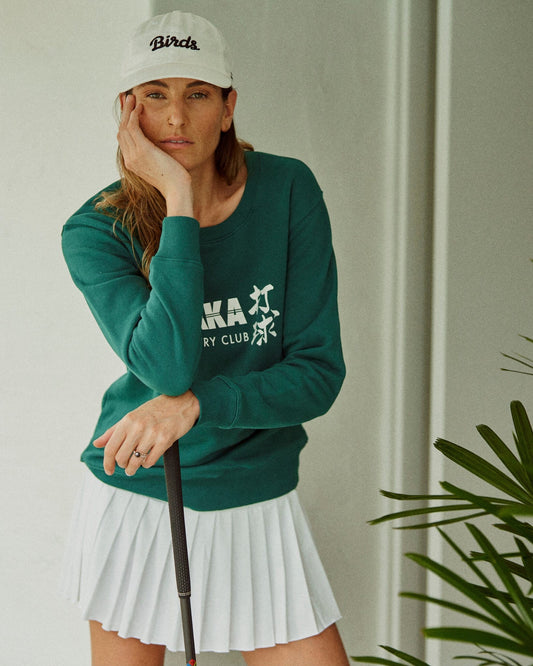 A woman wearing a white pleated skort paired with a dark green crewneck sweater and a white cap.