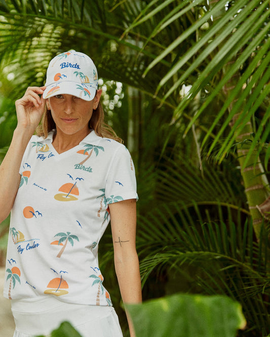 A woman wearing a white V-neck polo shirt and matching cap featuring a tropical beach print.