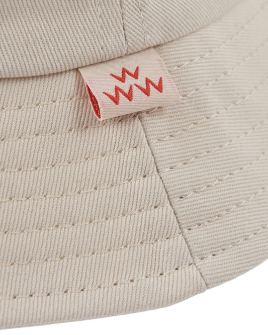 Detail of an off-white cotton twill bucket hat with a small red embroidered logo tag on the brim seam.