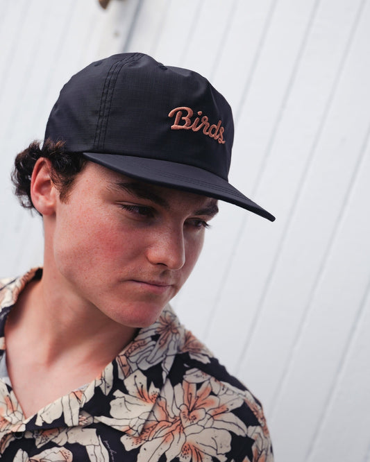 A man wearing a black snapback cap featuring embroidered pink script reading Birds on the front crown.