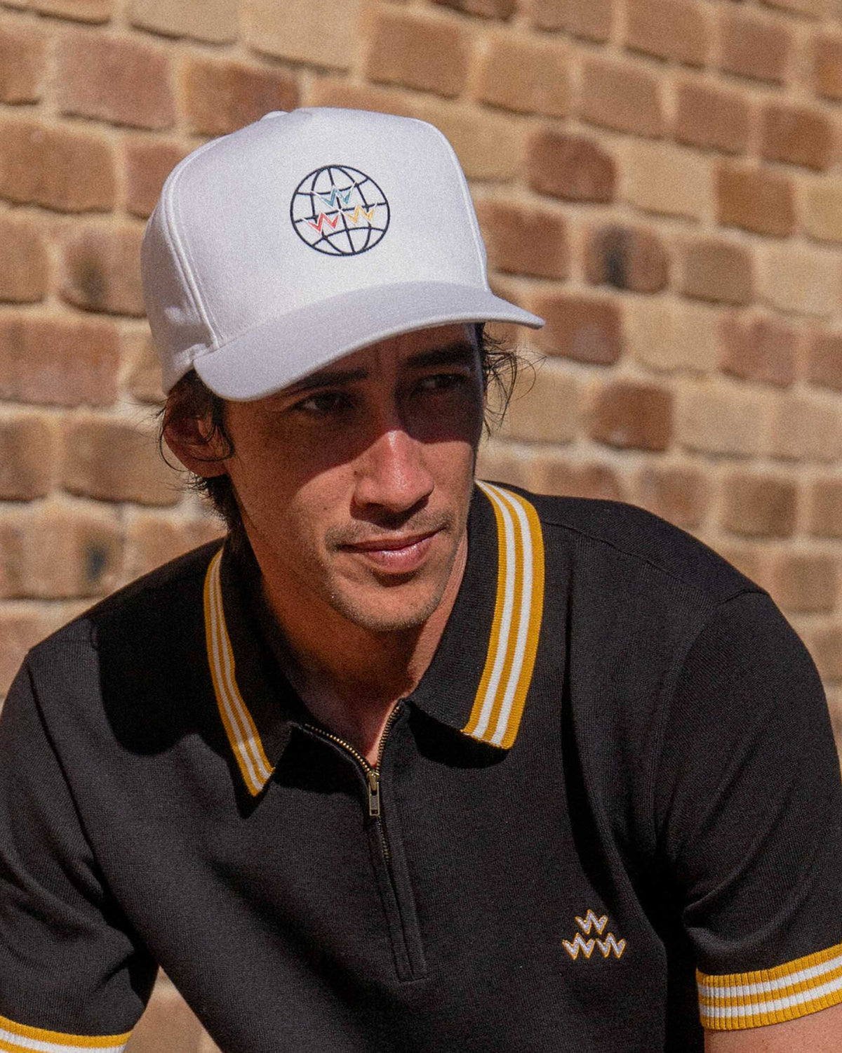 A man wearing a white snapback hat with a globe graphic, posed against a rustic brick wall outdoors.