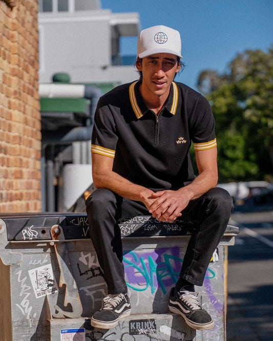 A man wearing a white snapback hat, black polo shirt with yellow collar trim, and black trousers.