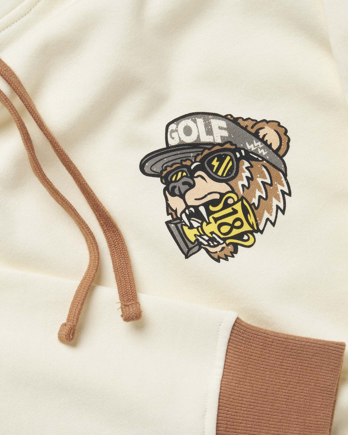 Detail of an off-white hoodie with a brown bear graphic, brown drawcords, and brown ribbing.