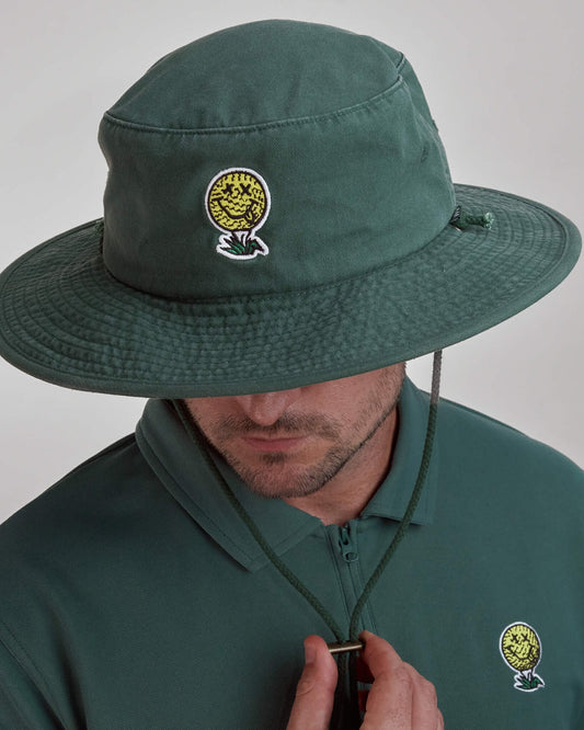 A man wearing a green boonie hat featuring a yellow smiley golf ball logo design on the front.