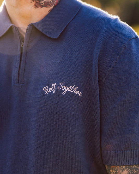 A person wearing a blue knit 1/4-zip polo shirt with white embroidered text on the chest.