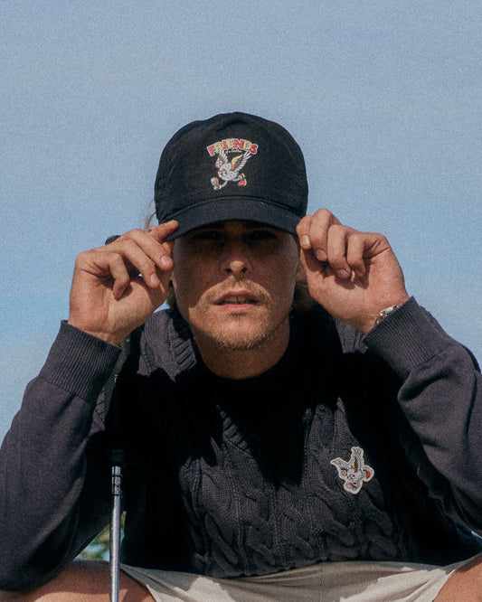 A man wearing a black snapback with a winged golf ball graphic on the front, and a dark knit sweater.