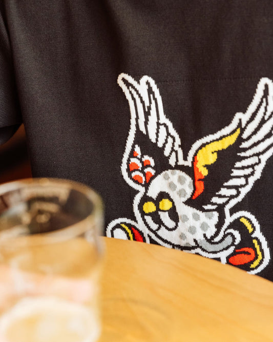 Detail of a black cotton sweater with a winged golf ball intarsia print worn at a table.
