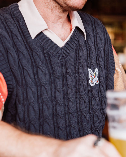 A person wearing a black cable knit V-neck vest featuring an embroidered winged golf ball badge.