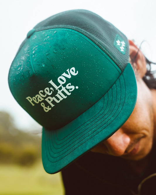 A man wearing a green trucker hat with a flat peak and white text reading "Peace, Love & Putts."