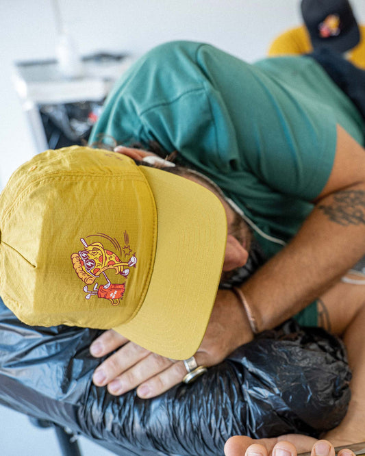 A man wearing a gold five-panel snapback cap featuring a pizza slice golf swing graphic on the side.