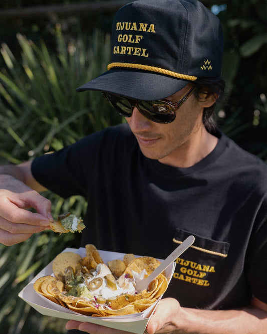 A man wearing a black snapback hat with a yellow rope and embroidered Tijuana Golf Cartel logo.