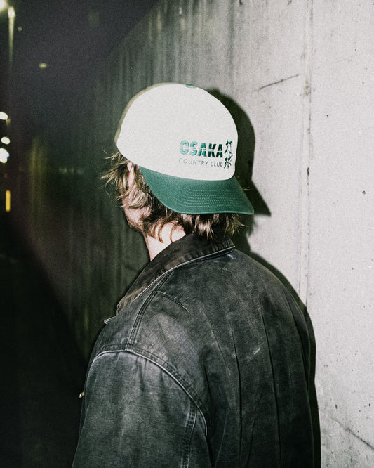 A man wearing a white snapback hat with a green brim and green Osaka Country Club text on the front.
