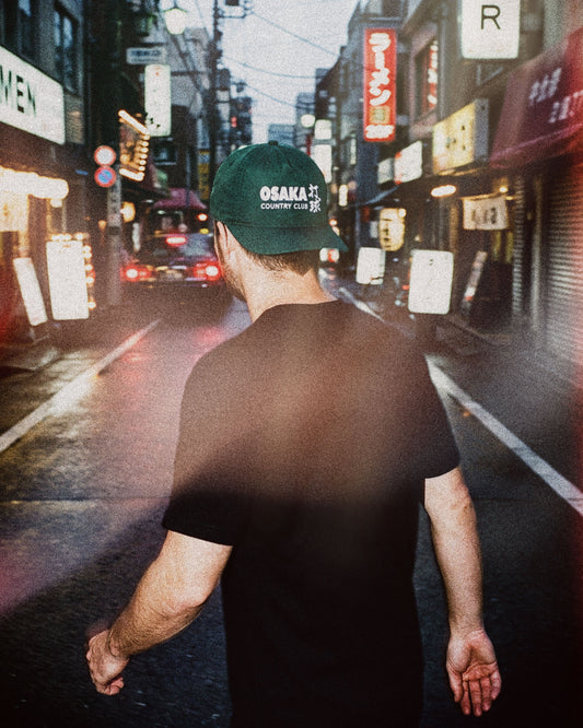 A man wearing an alpine green nylon snapback with white print, walking down a neon-lit narrow street.