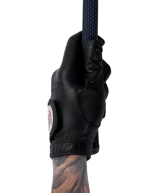 A hand wearing a black leather golf glove gripping a club, showing a circular patch on the wrist strap.