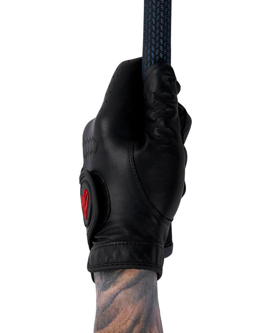A hand wearing a black leather golf glove with a red heart logo, gripping a textured golf club.