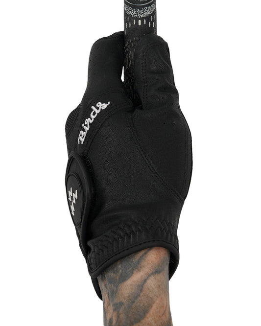 A hand wearing a black golf glove holding a club, featuring a white script script and a logo on the closure tab.
