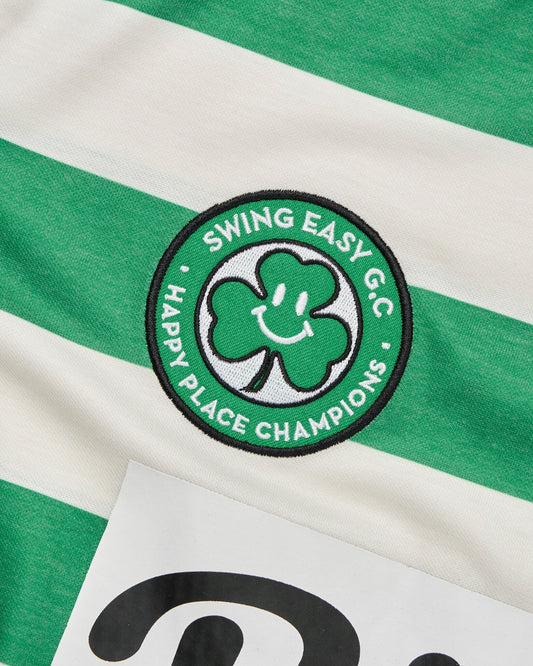 Detail of a green and white striped polo shirt featuring a circular woven patch with a smiley clover motif.