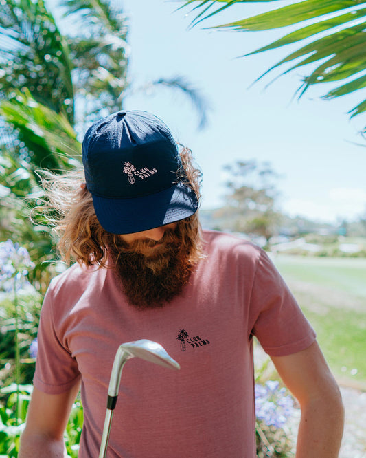 A bearded man wearing a navy snapback with a pale pink Club Palms print and a matching t-shirt.