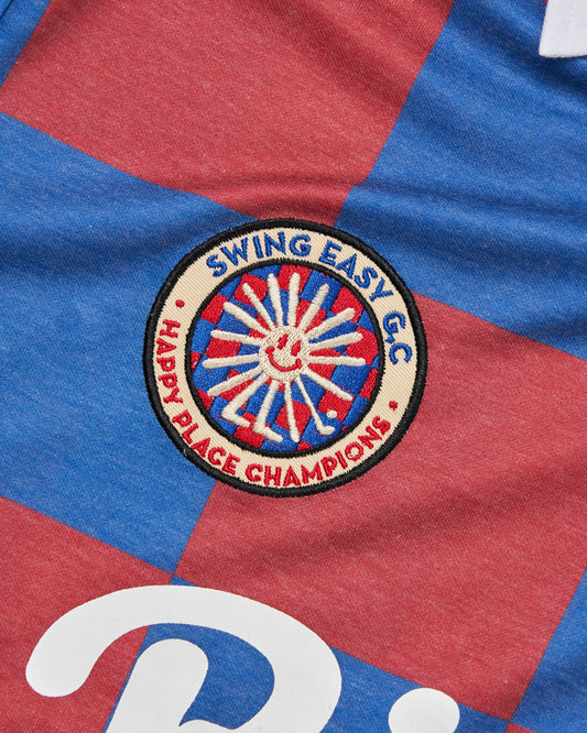 Detail of a burgundy and blue checkered polo shirt with a round patch and white chest graphic.