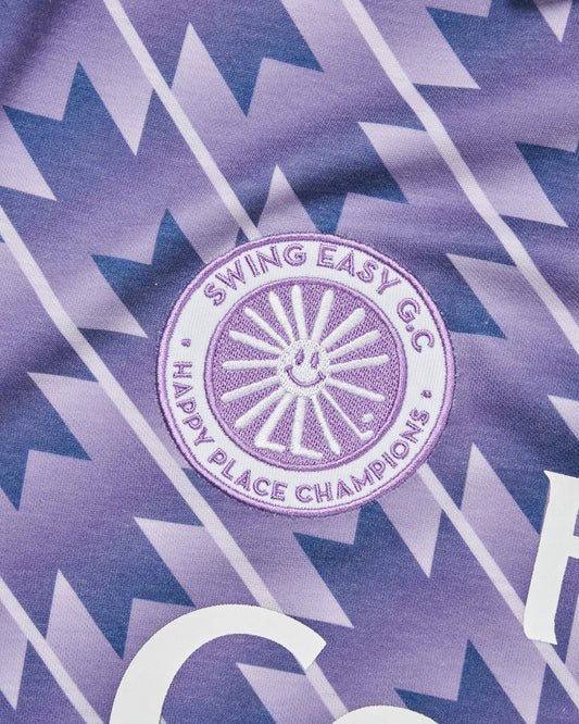 Detail of a purple haze polo shirt with a geometric pattern, round logo patch, and white chest print.