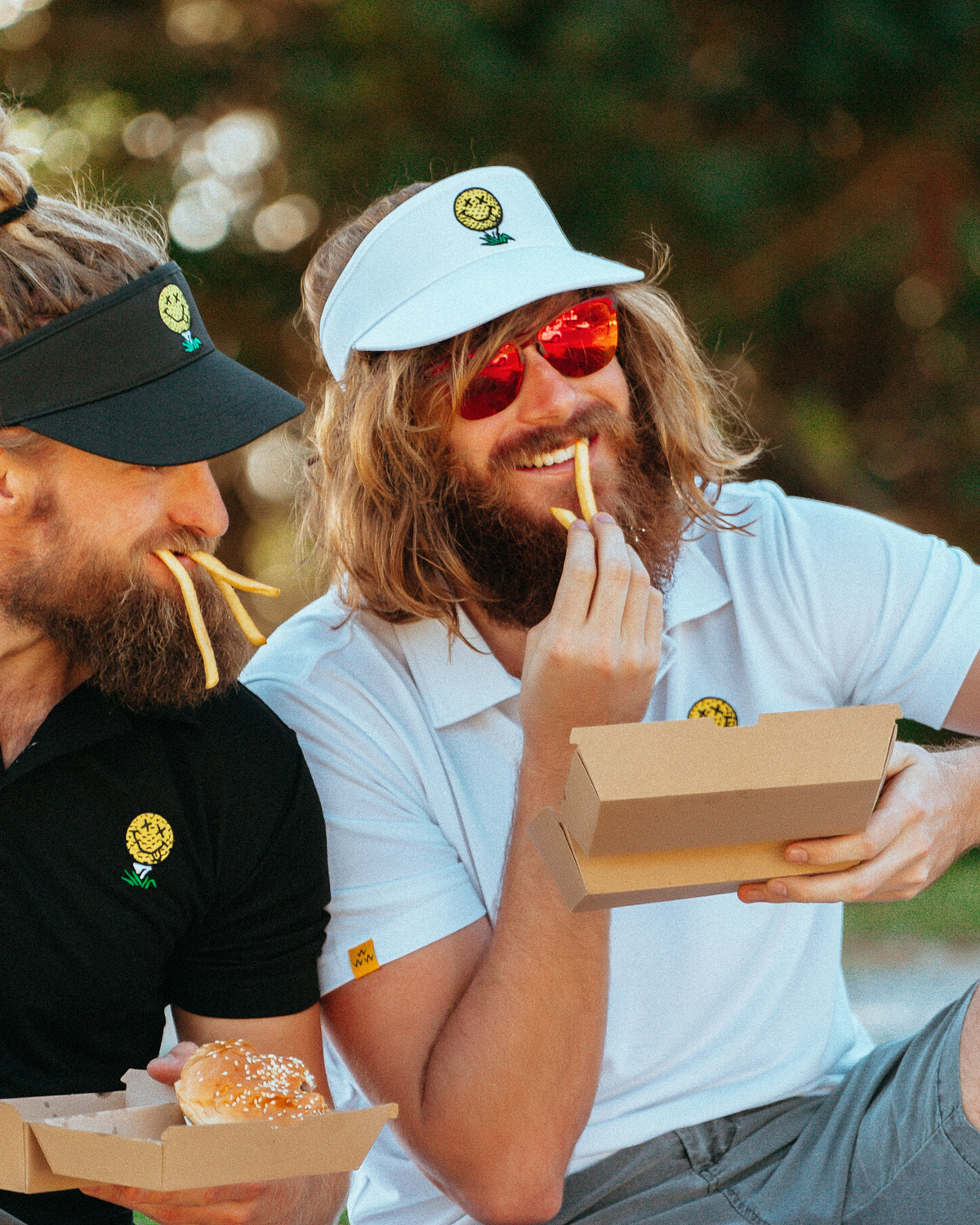 Two people wearing visors eat fast food; the man on the right wears a white visor with a smiley face.