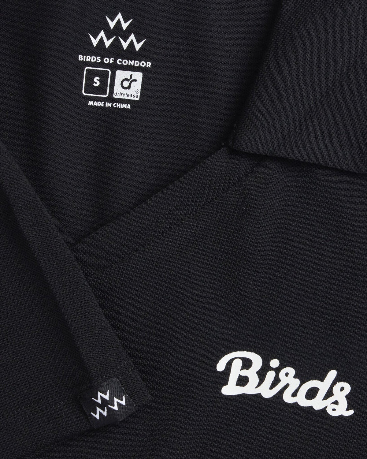Detail of a black pique polo shirt showing the collar, V-neck, interior brand label, and white script design.