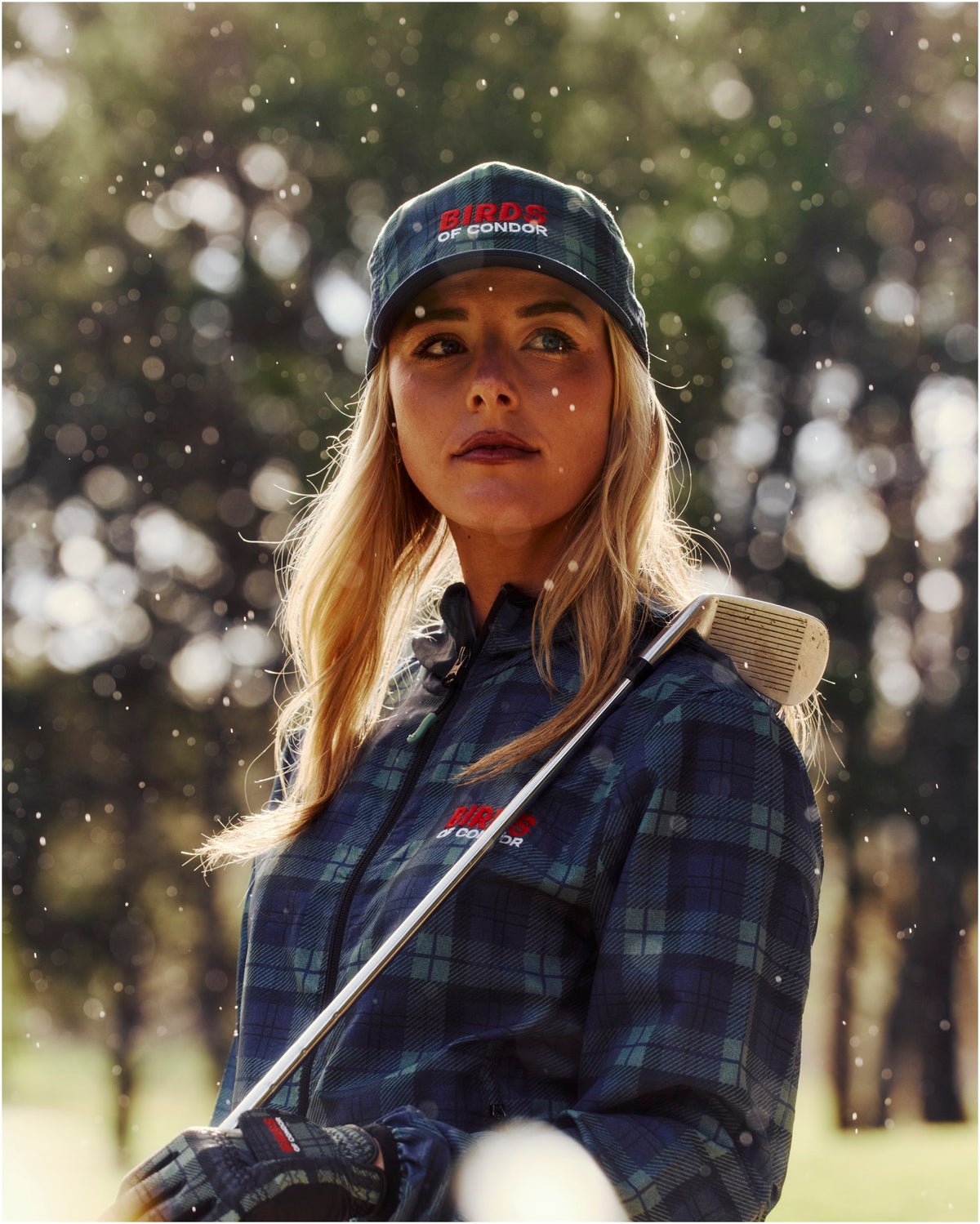 A woman wearing a blue and green plaid jacket and a matching cap, holding a golf club outdoors.