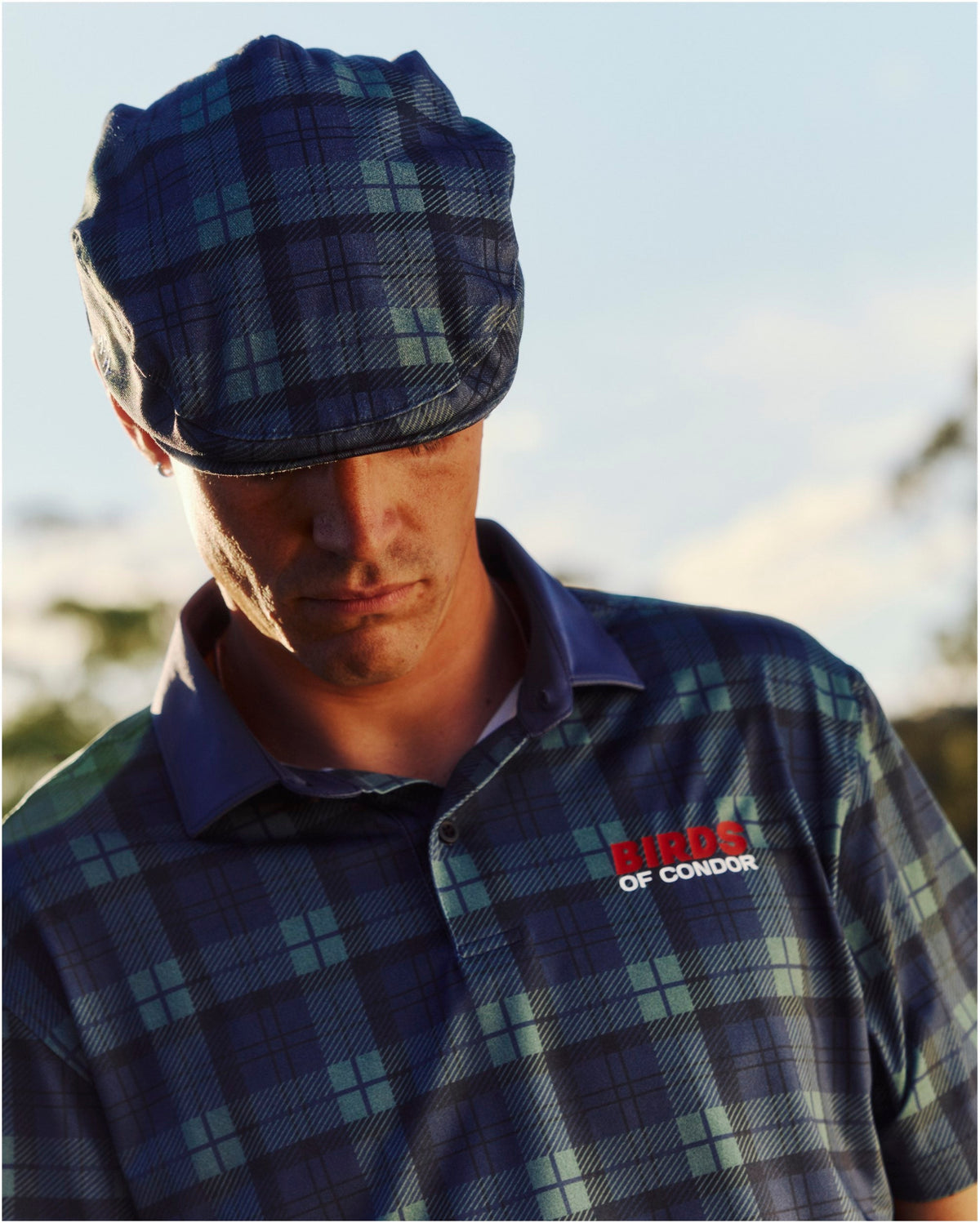 A man wearing a blue and green plaid polo shirt with a matching flat cap and a red and white chest logo.