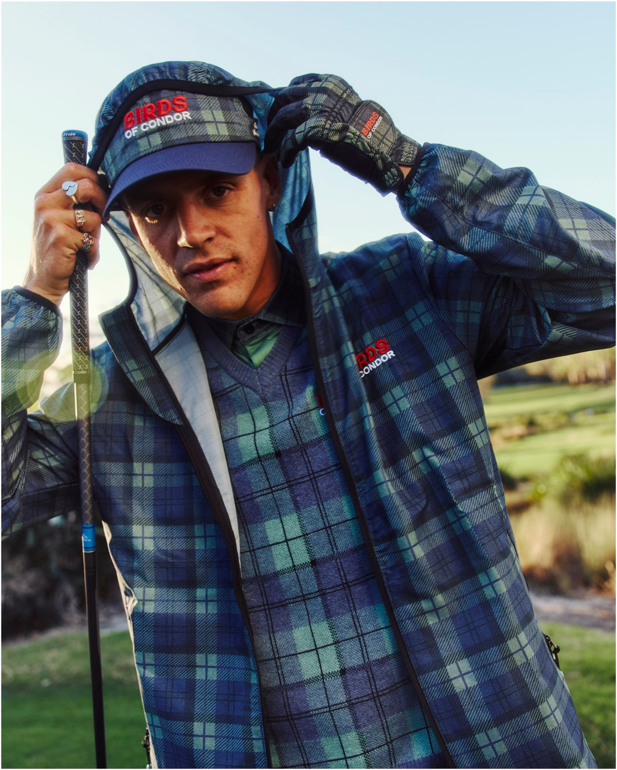 A man wearing a blue and green plaid shell jacket, matching visor, and a V-neck sweater on a golf course.