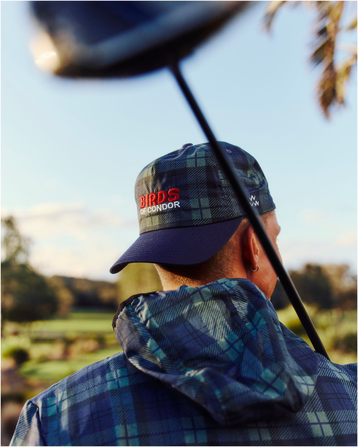 A person wearing a blue and green plaid jacket with a hood and a matching cap featuring embroidered red text.