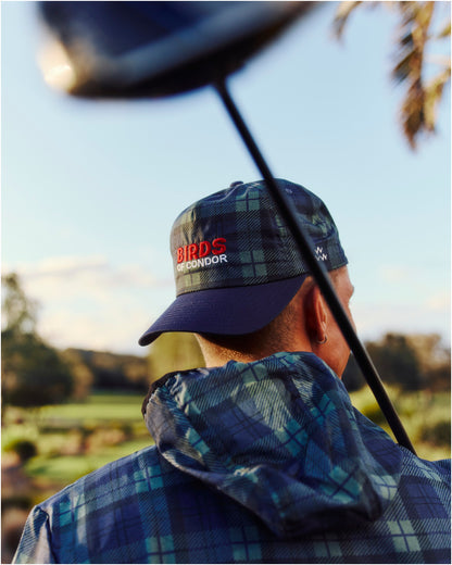 A person wearing a blue and green plaid jacket with a hood and a matching cap featuring embroidered red text.