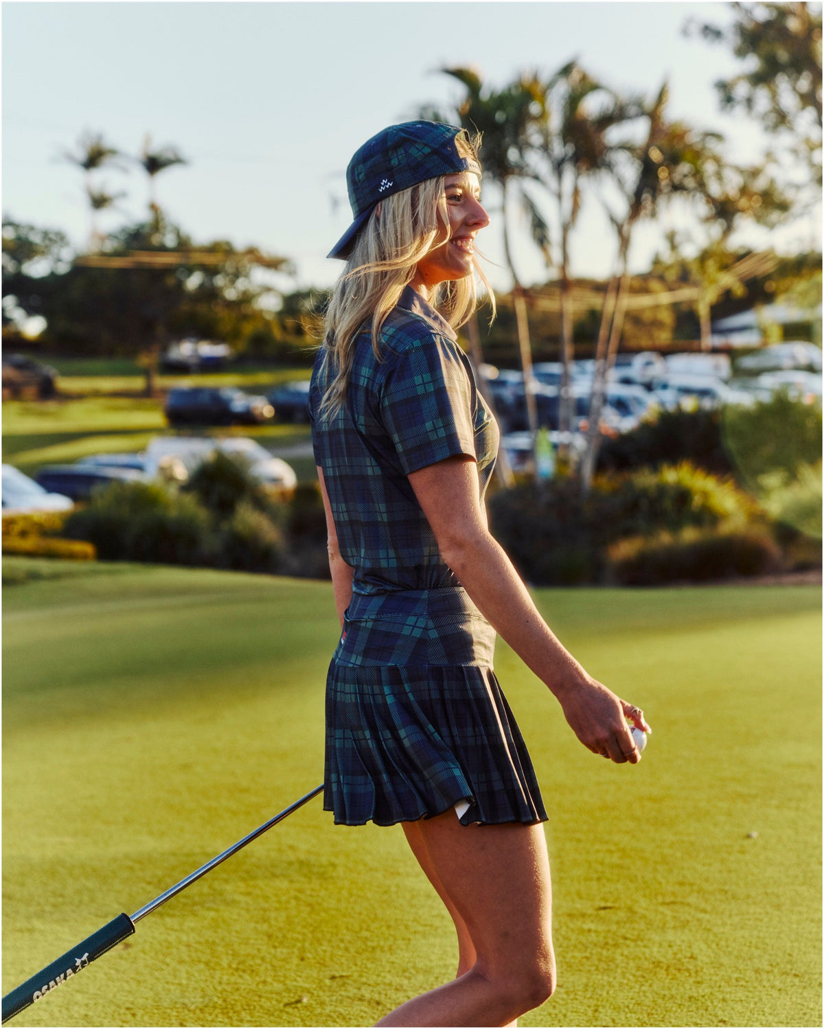 A woman wearing a blue and green plaid polo shirt and matching pleated skirt on a golf course.