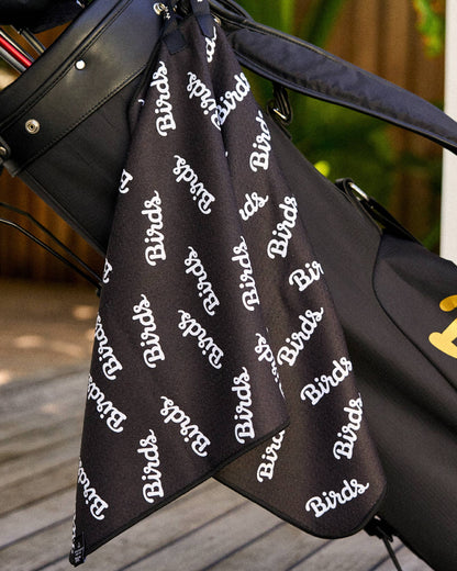 Black golf towel with an all-over white script Birds print, hanging from a black golf bag.
