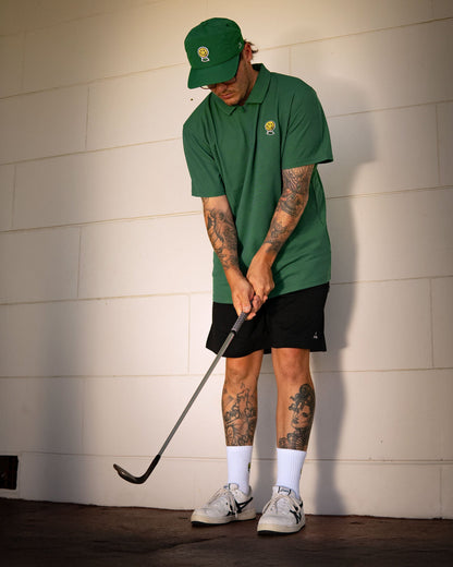 A man wearing a green polo shirt and matching cap, posing with a golf club against a white wall.