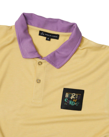 Detail of a tan polo shirt with a purple collar, two-button placket, and a square woven chest logo.