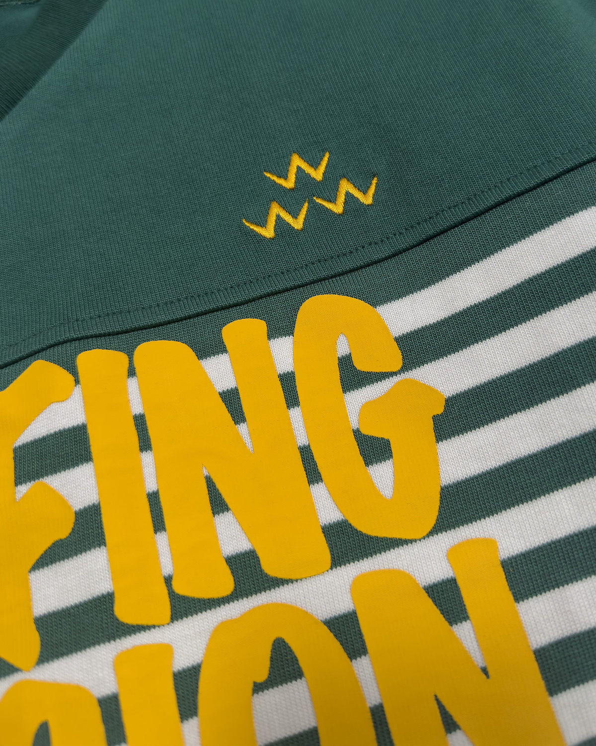 Detail of a green and white striped rugby sweater with yellow lettering and a three-w-motif.