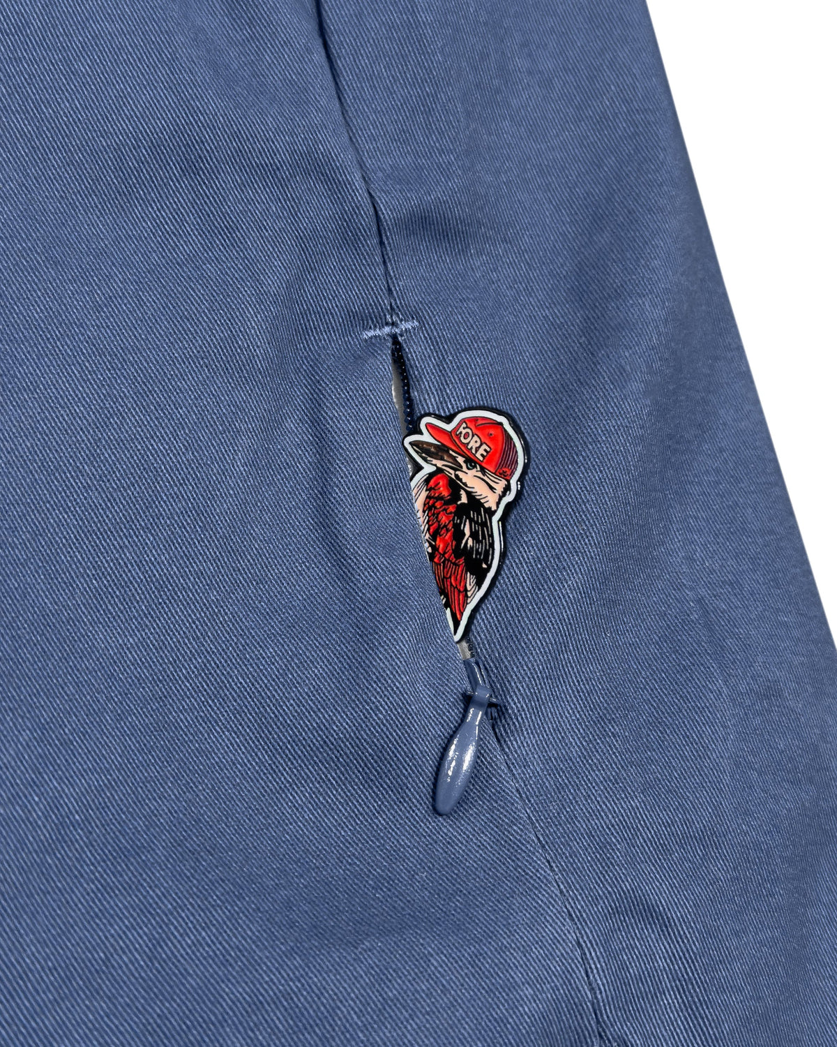 Detail of blue cotton pants showing a zipper pocket containing a bird graphic with a red hat.