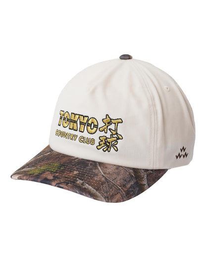 Natural snapback cap with a camouflage print brim and gold text embroidery on the front crown.