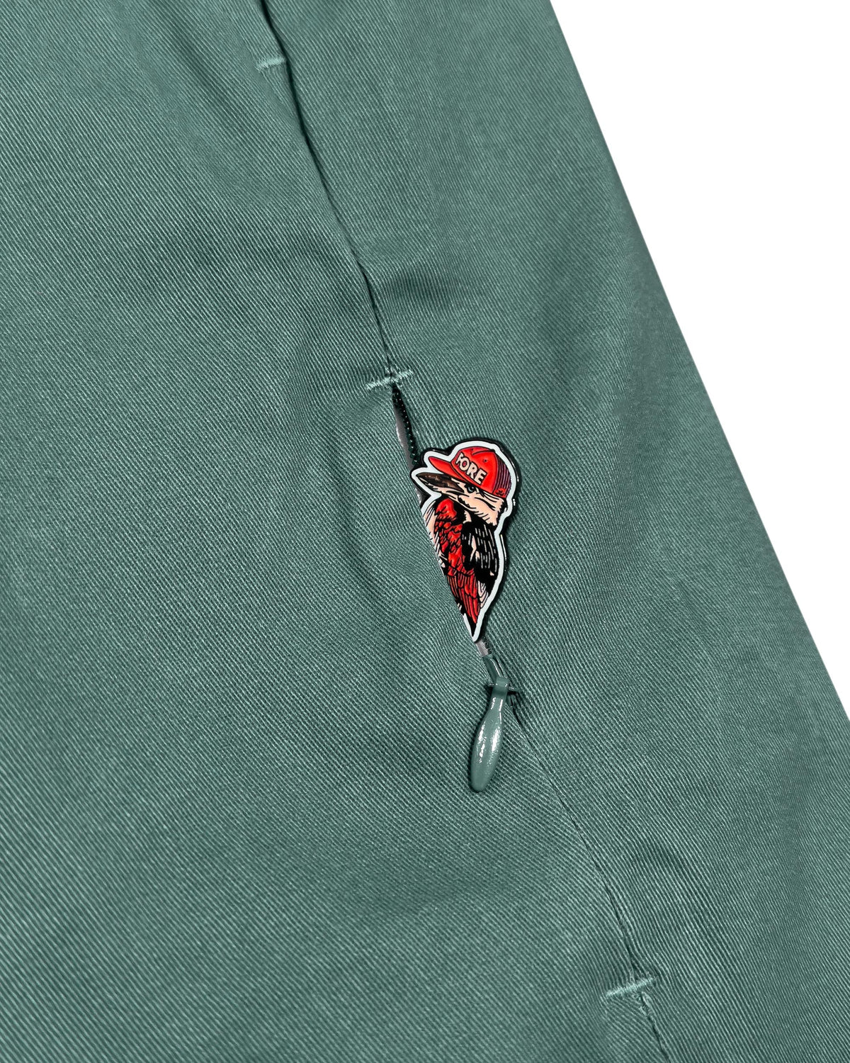 Detail of khaki green shorts featuring a concealed zip pocket with a printed bird motif pull.