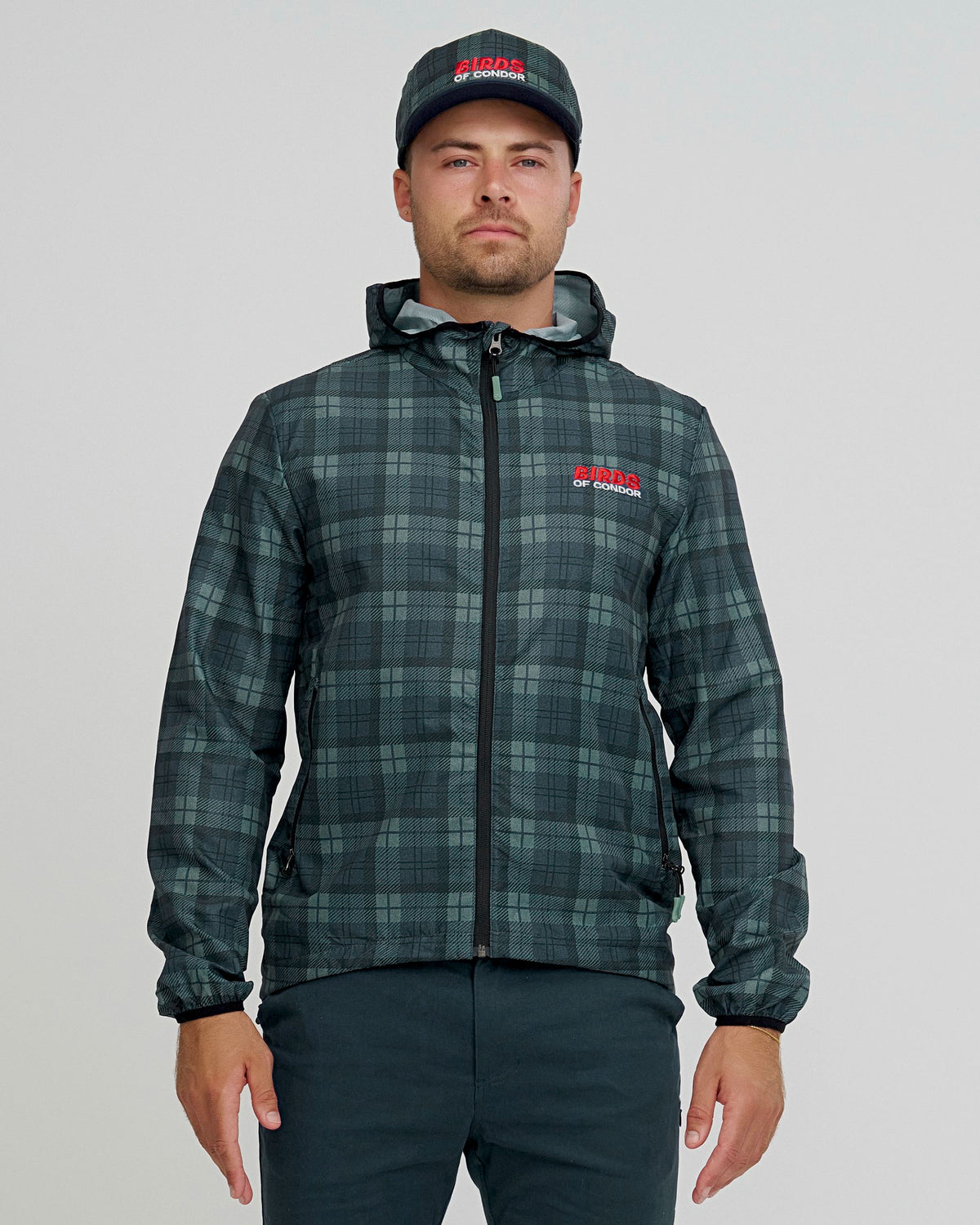 A man wearing a blue and green plaid jacket with a hood, a black zip front, and an embroidered red logo.