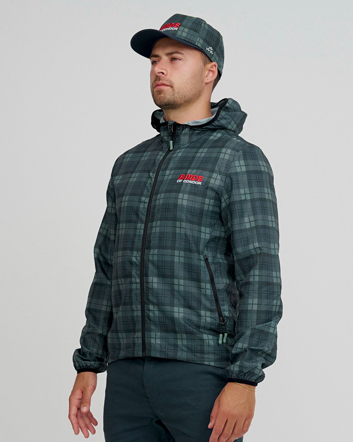 A man wearing a blue and green plaid shell jacket with an embroidered front logo, black zip, and matching cap.