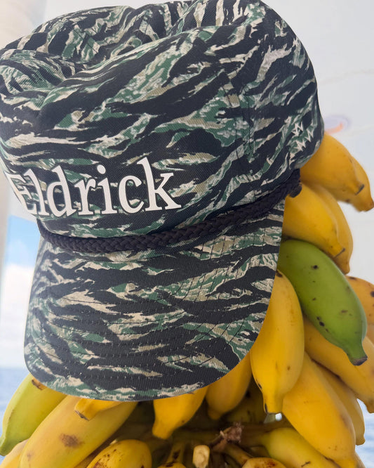 A close-up of a baseball cap with a tiger-stripe camouflage pattern in green, black, and beige, featuring the name 'Eldrick' in white text on the front. The cap is styled on top of a bunch of yellow and green bananas against a bright, overexposed background.