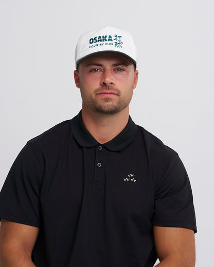 A man wearing a white snapback with a green brim and an Osaka Country Club graphic on the front.